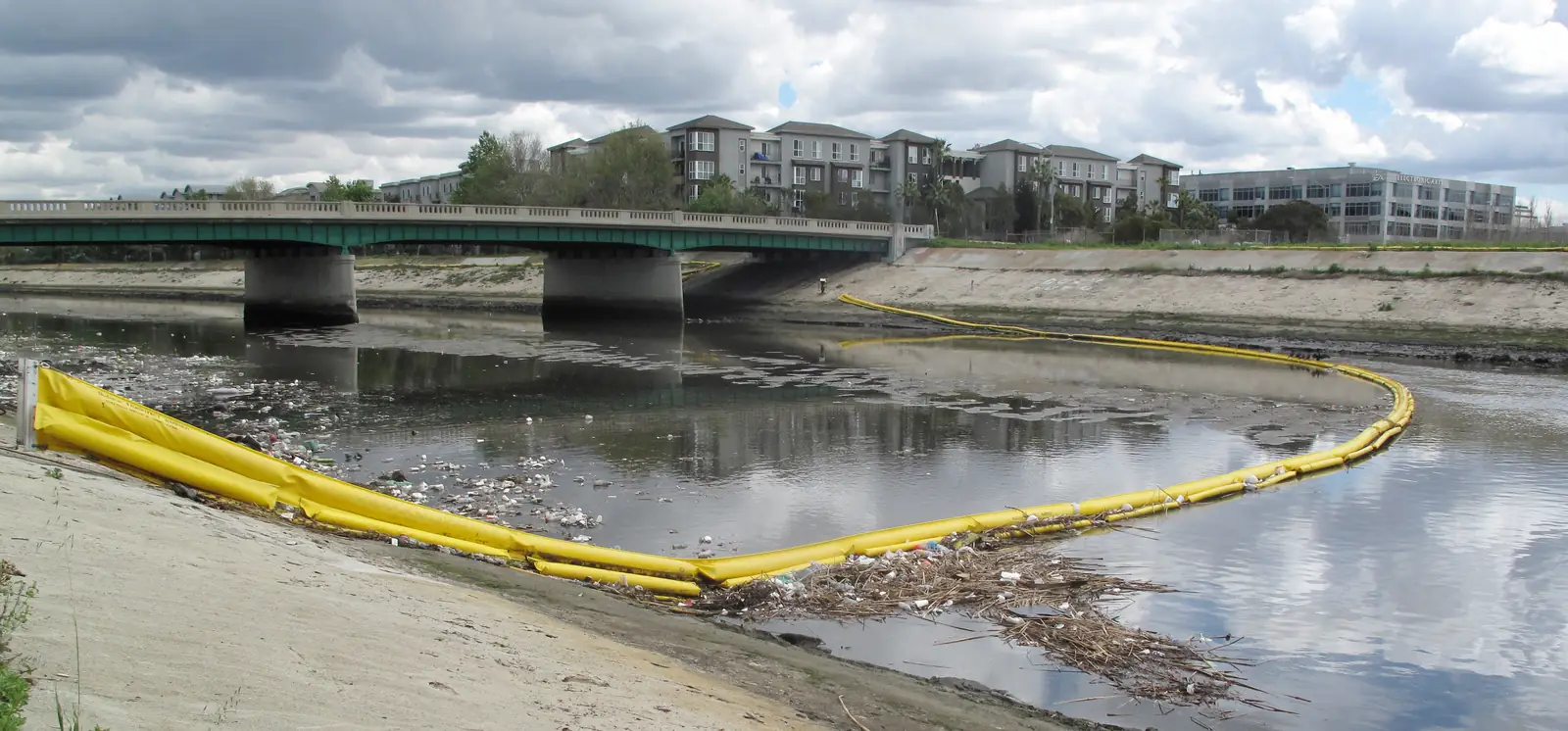 A yellow floating barrier curving across a concrete-lined creek, holding back floating trash under a bridge